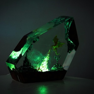 May include: A fantasy-themed resin sculpture featuring a green-glowing dragon breathing fire. The artwork is encased in a geometric, transparent block with a dark wooden base. A castle and other miniature figures are also visible.