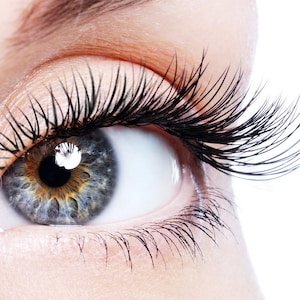 Op de afbeelding: Close-up van een menselijk oog met lange, zwarte wimpers. De iris is een mix van blauwe, grijze en bruine tinten, met een heldere witte reflectie. De huid rond het oog is glad en licht getint, tegen een witte achtergrond.