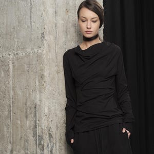 Könnte beinhalten: Schwarzes Langarm-Oberteil mit drapiertem Ausschnitt und passender schwarzer Hose mit Kordelzug. Das Outfit ist aus einem weichen Material gefertigt. Das Model trägt schwarze Schuhe. Der Hintergrund umfasst eine Betonmauer und schwarze Vorhänge.