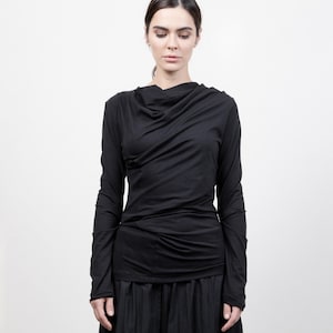 Peut inclure: Un chemisier noir à manches longues avec un col drapé et une jupe noire à taille froncée. La jupe est asymétrique et tombe au niveau du genou d'un côté et de la cheville de l'autre.