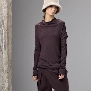 Könnte beinhalten: Ein dunkelviolettes Langarmshirt mit drapiertem Wasserfallausschnitt und eine passende dunkelviolette weite Hose mit Wickeloptik vorne. Das Outfit wird mit einem beigen, flauschigen Fischerhut und schwarzen Schuhen zu einem monochromen Look kombiniert.