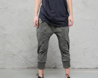 May include: A black baseball cap, a dark navy sleeveless top with a draped cowl neckline, olive green cropped jogger pants with ribbed cuffs, and black athletic sneakers. The outfit features a layered aesthetic with a relaxed, loose-fitting silhouette.