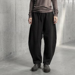 Op de afbeelding: Een donkergrijze bucket hat, een taupekleurig shirt met lange mouwen, een hoge halslijn en zichtbare stiksels, en een losvallende zwarte broek met een trekkoord in de taille. De outfit wordt afgemaakt met donkergrijze leren schoenen.