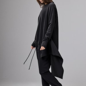 Könnte beinhalten: Eine schwarze Langarmtunika mit asymmetrischem Saum und dünnen Kordelzügen, kombiniert mit einer passenden schwarzen Jogginghose und schwarzen Lederstiefeletten mit seitlichen Reißverschlüssen. Das Outfit ist einfarbig und weit geschnitten.