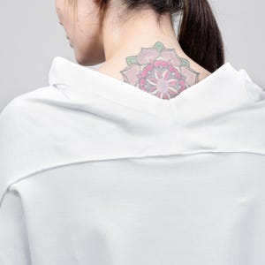 May include: A white, short-sleeved, hooded shirt with a loose fit. The shirt has a simple design and is made of a soft, comfortable fabric. The model is wearing the shirt and has a pink flower tattoo on their neck.