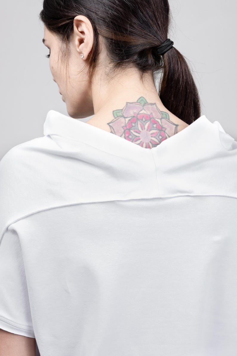 May include: A white, short-sleeved, hooded shirt with a loose fit. The shirt has a simple design and is made of a soft, comfortable fabric. The model is wearing the shirt and has a pink flower tattoo on their neck.