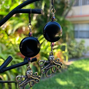 May include: Black onyx gemstone earrings with a dangling octopus charm. The earrings are made with brass and have a black onyx gemstone.