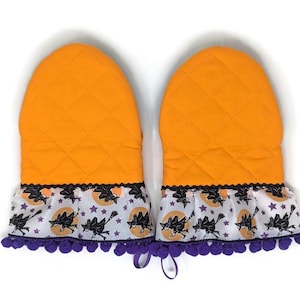 May include: A pair of orange quilted oven mitts with a decorative ruffle. The ruffle features a black witch on a broomstick design with purple stars and a purple pom-pom trim. The mitts have a loop for hanging.