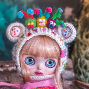 May include: A doll wearing a handmade white knit hat with colorful embellishments and ear-shaped details. The hat is adorned with whimsical, cartoonish faces and colorful accents. The doll has large blue eyes and blonde hair.