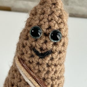 May include: A brown crocheted stuffed toy with a smiling face and large blue eyes. The toy is wrapped in a brown, white, and beige yarn.