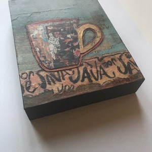 May include: A mixed media artwork featuring a brown and gold coffee cup with a textured background. The text "Java Java" is written in black paint on the bottom of the artwork. The artist's signature is visible in the bottom right corner.
