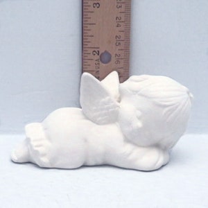 Może przedstawiać: Biała ceramiczna figurka śpiącego anioła z skrzydłami. Anioł leży na boku z głową opartą na ramieniu. Figurka ma około 13 cm długości.
