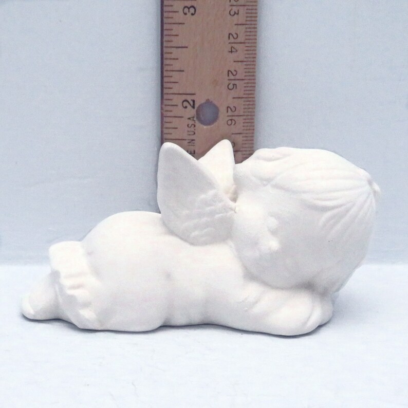 Może przedstawiać: Biała ceramiczna figurka śpiącego anioła z skrzydłami. Anioł leży na boku z głową opartą na ramieniu. Figurka ma około 13 cm długości.
