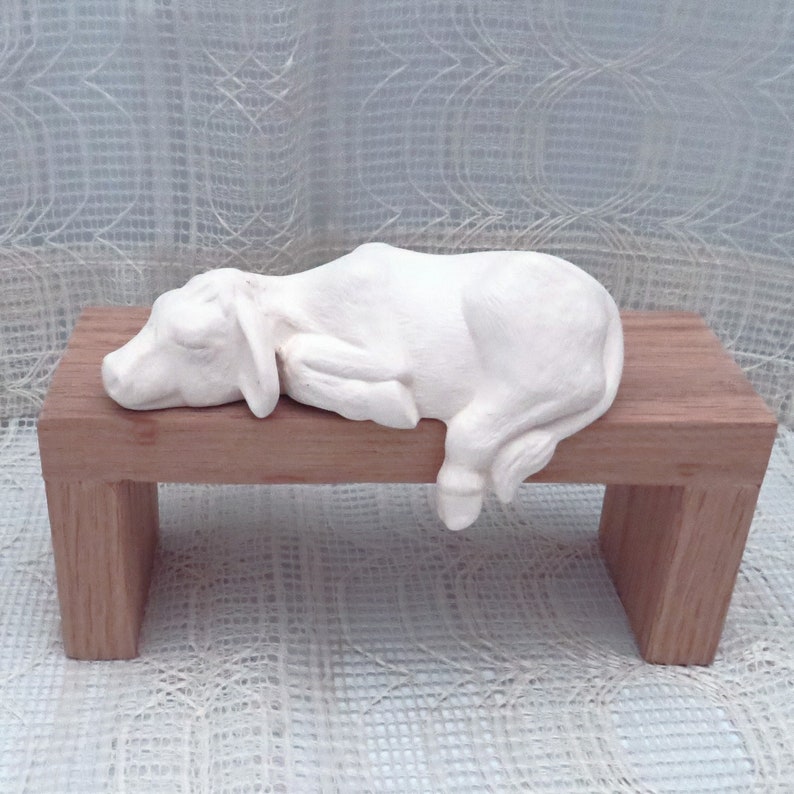 May include: A white ceramic sculpture of a cow lying down on a wooden bench. The cow is in a sleeping position with its head resting on the bench.