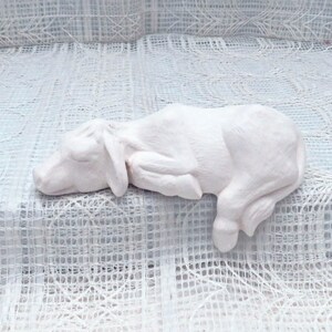 May include: White ceramic figurine of a sleeping cow, lying on its side with its head resting on its front legs. The figurine is unpainted and has a smooth, matte finish.