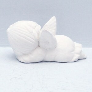 Może przedstawiać: Biała ceramiczna figurka anioła leżąca na boku z rozłożonymi skrzydłami za sobą. Anioł ma prosty, dziecięcy design i idealnie nadaje się do dodania odrobiny fantazji do każdego wystroju domu.