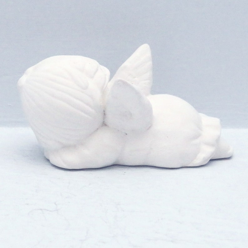 Może przedstawiać: Biała ceramiczna figurka anioła leżąca na boku z rozłożonymi skrzydłami za sobą. Anioł ma prosty, dziecięcy design i idealnie nadaje się do dodania odrobiny fantazji do każdego wystroju domu.
