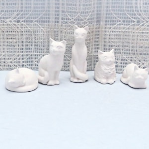 May include: Five white ceramic cat figurines. The cats are in various poses, including sitting, lying down, and curled up. The figurines are all made of the same white ceramic and have a smooth, matte finish.