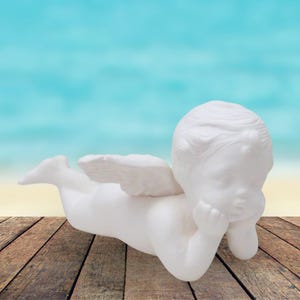 May include: White ceramic angel figurine with wings, lying on its stomach with its head resting on its hand. The angel has a cherubic face and is painted in a matte white finish.