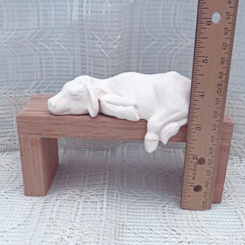 May include: White ceramic figurine of a sleeping calf lying on a wooden bench. The bench is about 7 inches long.
