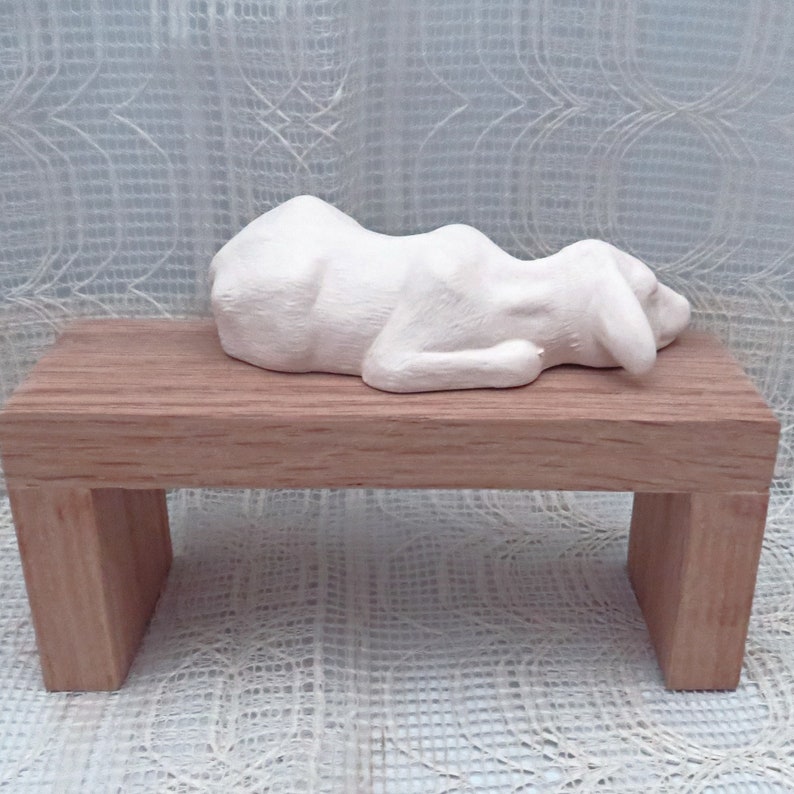 May include: White ceramic sculpture of a dog lying down on a wooden bench. The dog is in a sleeping position with its head resting on its paws.