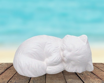 May include: White ceramic figurine of a cat sleeping on a wooden surface. The cat is curled up in a ball with its head resting on its paws.