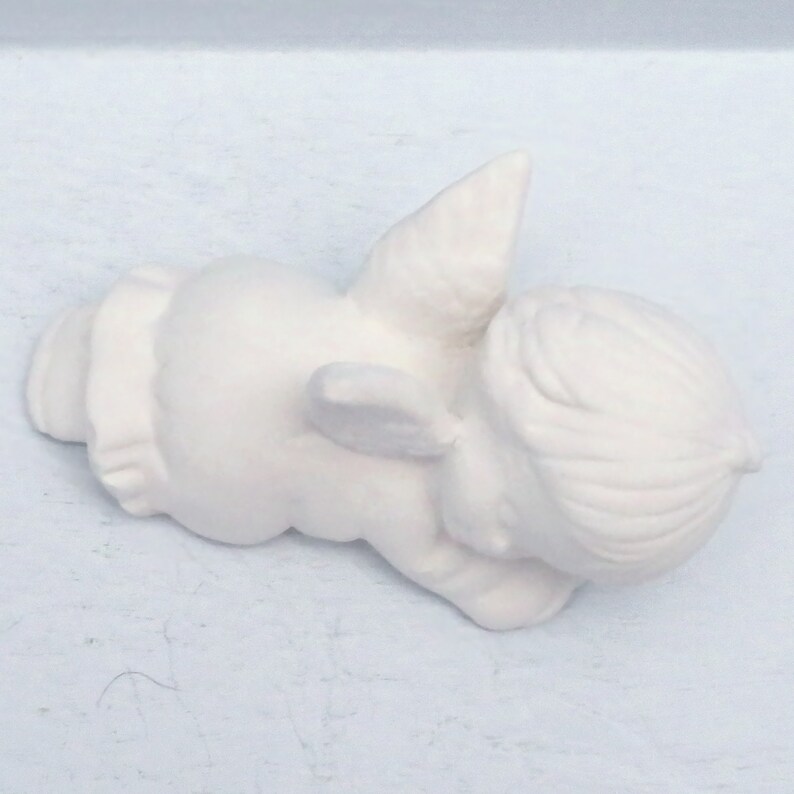 Może przedstawiać: Biała ceramiczna figurka śpiącego anioła z skrzydłami. Anioł leży na boku z głową opartą o ramię.