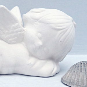 Może przedstawiać: Biała ceramiczna figurka anioła z skrzydłami, leżąca na boku z głową opartą o ramię. Anioł ma prosty, delikatny design.