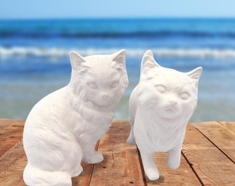 May include: Two white ceramic cat figurines, one sitting and one standing, on a wooden surface. The background is a blurred image of a beach.