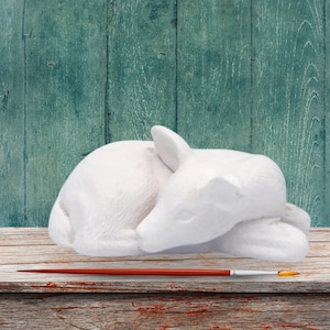 May include: A white ceramic deer figurine in a sleeping position. The deer is lying on a wooden surface with a paintbrush in front of it. The background is a teal-colored wooden wall.
