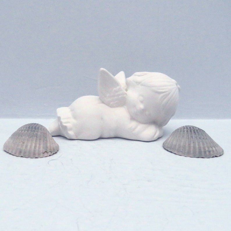 Może przedstawiać: Biała ceramiczna figurka anioła z skrzydłami, leżąca na boku z głową opartą na ramieniu. Anioł ma słodki, niewinny wyraz twarzy. Figurka jest otoczona dwoma szarymi muszelkami.