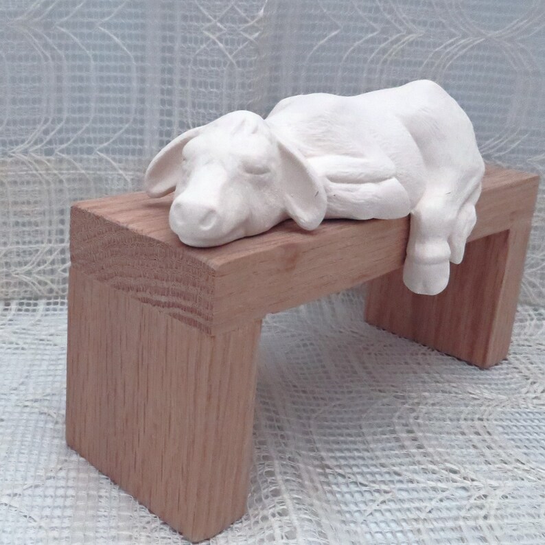 May include: White ceramic figurine of a sleeping cow lying on a wooden bench. The bench is made of light brown wood and has a simple design.