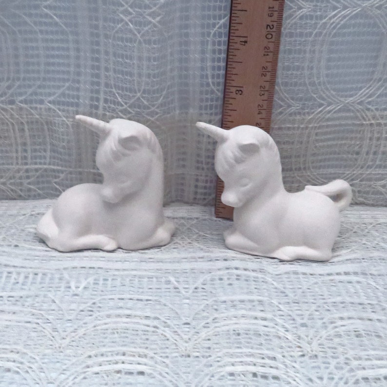 Op de afbeelding: Twee witte keramische eenhoornbeeldjes, elk ongeveer 10 cm hoog, liggend op een wit kant oppervlak.