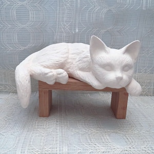 May include: White ceramic cat figurine lying down on a wooden bench. The cat has a detailed face and fur texture.
