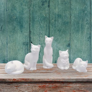 May include: A set of five white ceramic cat figurines in various poses, including one sleeping, one sitting upright, one standing tall, one sitting with a fluffy texture, and one lying down, all featuring a smooth, matte, unpainted white finish.