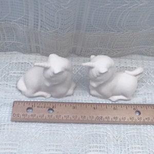 Op de afbeelding: Twee witte keramische eenhoornbeeldjes, elk ongeveer 7,5 cm lang, liggend met hun hoofd naar de zijkant gedraaid. De eenhoorns kijken elkaar aan.