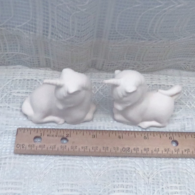 Op de afbeelding: Twee witte keramische eenhoornbeeldjes, elk ongeveer 7,5 cm lang, liggend met hun hoofd naar de zijkant gedraaid. De eenhoorns kijken elkaar aan.
