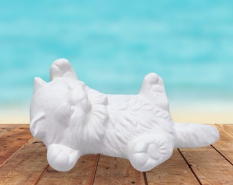 May include: White ceramic cat figurine lying on its back with its paws in the air. The figurine is on a wooden surface.