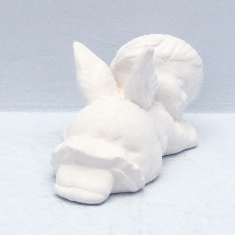 Może przedstawiać: Biała ceramiczna figurka śpiącego anioła z skrzydłami. Anioł leży na boku z głową opartą na ramieniu.