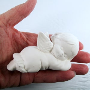 Może przedstawiać: Biała ceramiczna figurka anioła z skrzydłami, leżąca na boku z głową opartą o ramię. Anioł ma słodki, niewinny wyraz twarzy.