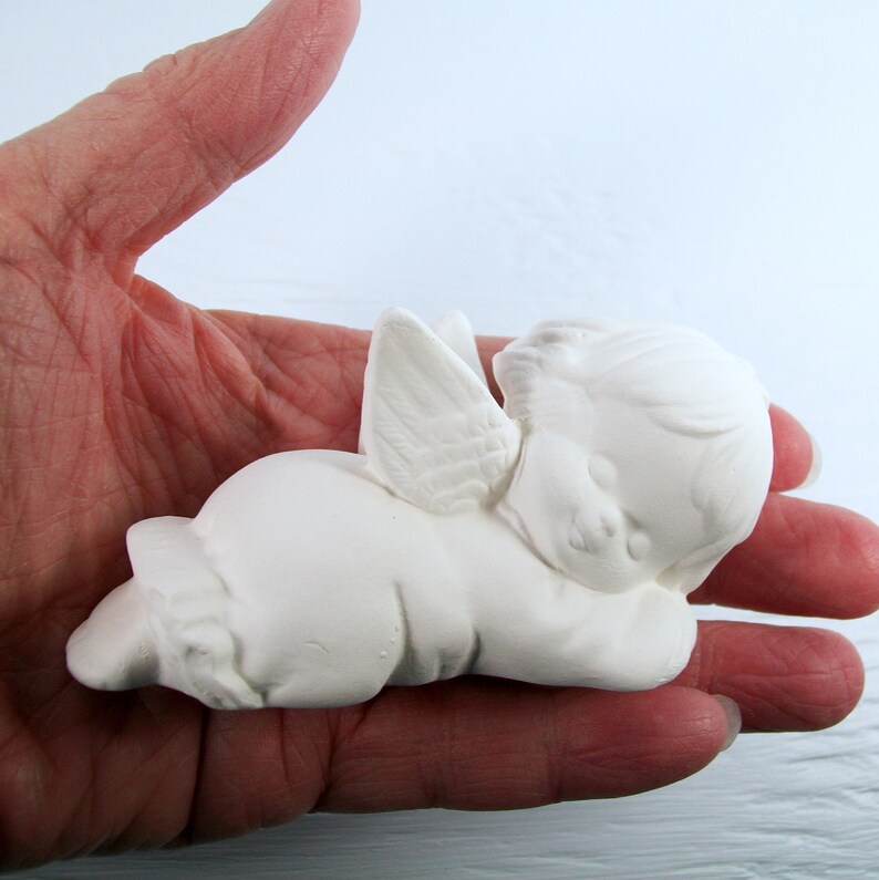 Może przedstawiać: Biała ceramiczna figurka anioła z skrzydłami, leżąca na boku z głową opartą o ramię. Anioł ma słodki, niewinny wyraz twarzy.