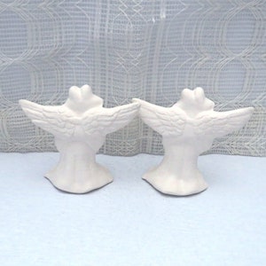 Back view of paintable ceramic frog angels showing the outstretched wings and tunics. They are on a pale blue table and in front of a lacy curtain.