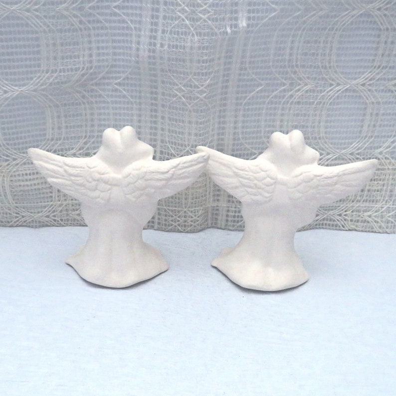 Back view of paintable ceramic frog angels showing the outstretched wings and tunics. They are on a pale blue table and in front of a lacy curtain.