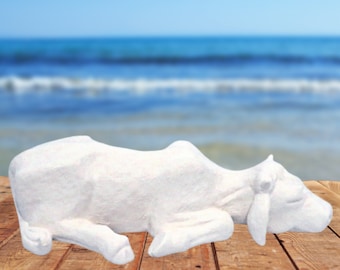 May include: White ceramic sculpture of a cow lying down on a wooden surface. The cow is in a relaxed pose with its head resting on the surface. The background is a blurred image of a blue ocean.