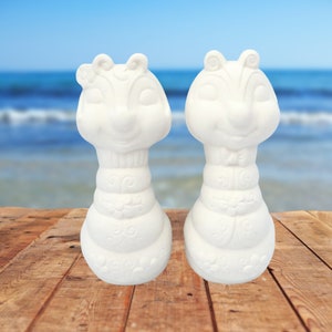 May include: Two white ceramic figurines of chipmunks, each with a detailed design and a whimsical expression. The chipmunks are standing on a wooden surface.