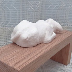 May include: White ceramic sculpture of a sleeping rabbit lying on a wooden bench.