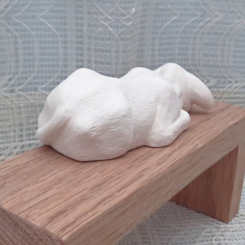 May include: White ceramic sculpture of a sleeping rabbit lying on a wooden bench.