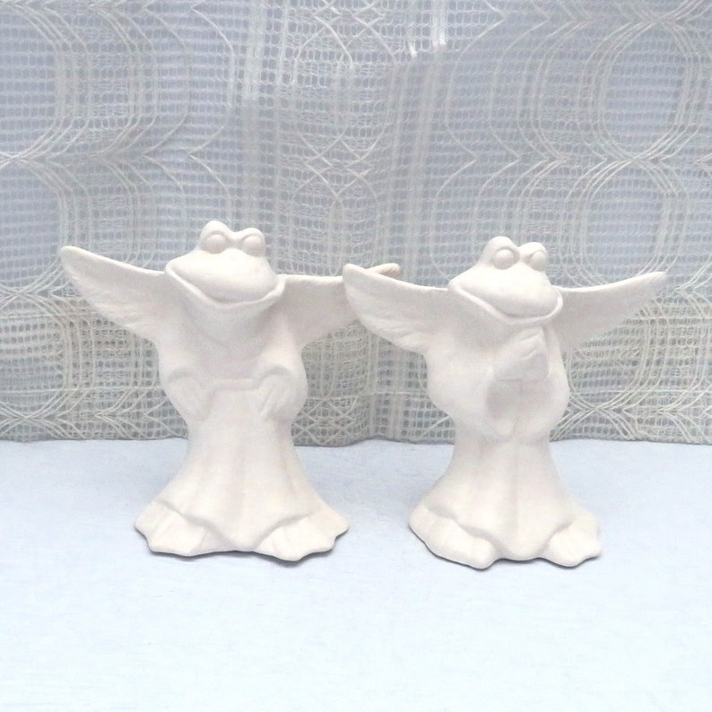 2 unpainted ceramic frog angels standing on a pale blue background with an ecru lacy curtain behind them.