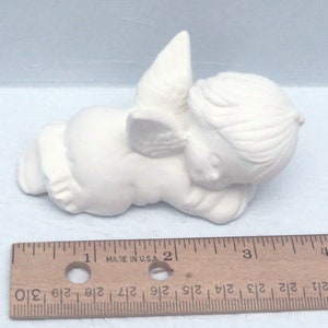 Może przedstawiać: Biała ceramiczna figurka anioła z skrzydłami, leżąca na boku z głową opartą o ramię. Anioł ma około 5 cm długości.
