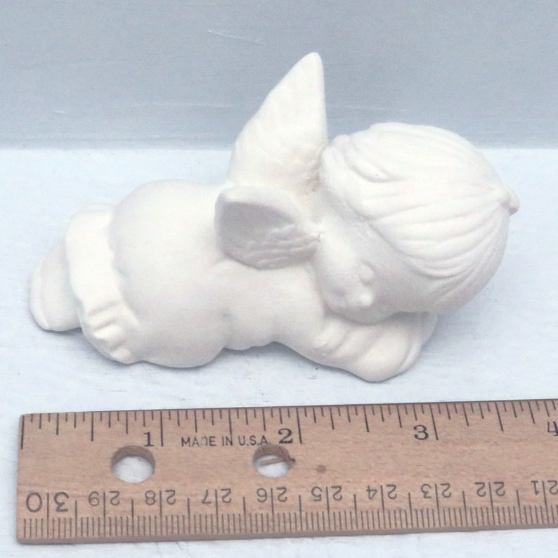 Może przedstawiać: Biała ceramiczna figurka anioła z skrzydłami, leżąca na boku z głową opartą o ramię. Anioł ma około 5 cm długości.